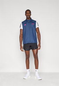 Navy sleeveless jacket with a full zipper, pink accents, and white short-sleeved shirt underneath, paired with black shorts and white sneakers.