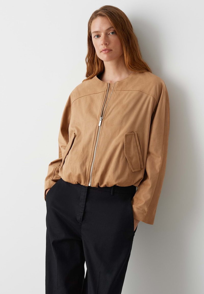 Giubbotto bomber beige in materiale morbido, con zip frontale, scollatura rotonda e taschino. Abbinato a pantaloni neri.