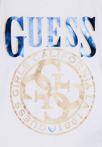 Valge T-särk, millel on suur sinine "GUESS" tekst ja kuldne ringikujuline logo, millel on tekst "GIRLS CALIFORNIA LA 1981" ning väike sinine südame detail.