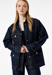 Jeansowa kurtka w ciemnoniebieskim kolorze, o luźnym kroju, z napami, dwoma kieszeniami na piersi i kontrastowym przeszyciem. Miękka w dotyku, z długimi rękawami.