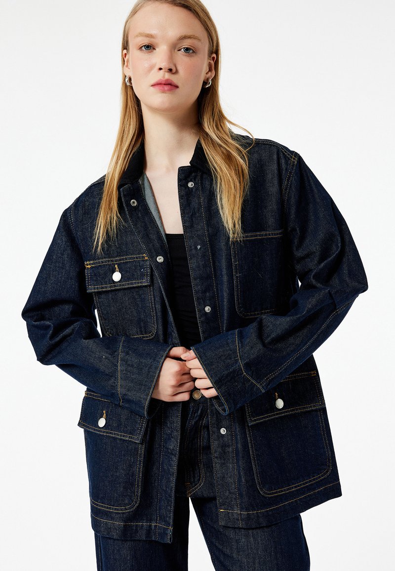 Jeansowa kurtka w ciemnoniebieskim kolorze, o luźnym kroju, z napami, dwoma kieszeniami na piersi i kontrastowym przeszyciem. Miękka w dotyku, z długimi rękawami.