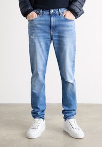 Ljust blå denimjeans med ett lätt slitna knä, rak passform, fem fickor och metallbeslag. Kombinerat med vita sneakers.