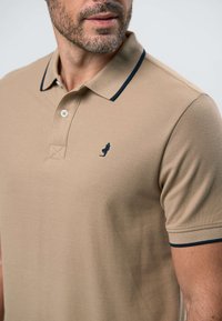 Bărbat purtând un tricou polo bej cu margini închise la culoare la guler și mâneci, cu un mic logo brodat albastru închis pe piept.