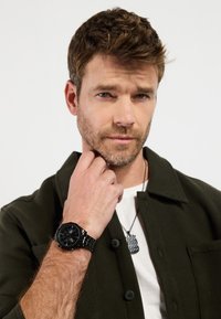 Schwarze Uhr mit rundem Zifferblatt und Metallarmband, am Handgelenk getragen. Mann in einer dunkelgrünen Jacke über einem weißen Hemd, mit einer silbernen Anhänger-Halskette.