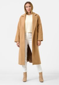 Cappotto lungo beige con spalle scese, tessuto testurizzato e maniche larghe, indossato sopra un maglione di maglia bianco e pantaloni bianchi, abbinato a stivali alla caviglia beige.