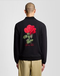 Crni dukserica s velikim vezom crvene ruže na leđima, sa zelenim lišćem i crvenim natpisom "Lyle and Scott". Uski kroj, rebrasti ovratnik.