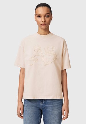 Kvinde med mørkt hår trukket tilbage iført en beige oversized t-shirt med hævede AS-bogstaver og blå jeans mod en ensfarvet baggrund.