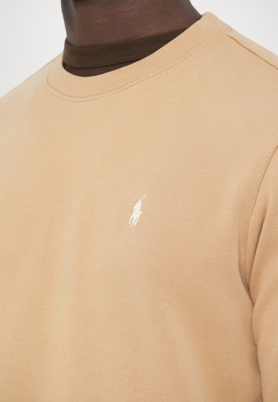 Bež pulover z okroglim izrezom, teksturiranim tkanjem in majhnim belim logotipom na prsih. Nošen čez temno rjavo srajco.