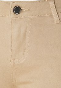 Pantalon beige avec une texture lisse, présentant une poche avant, des coutures contrastantes et un bouton en métal à la taille pour la fermeture.