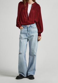 Röd, plisserad blus med knäppning fram, kombinerad med ljusblå högmidjade vida jeans med ett hål vid knät. Svarta kraftiga skor.
