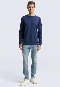 Navy blue sweatshirt with long sleeves, crew neck, and ribbed cuffs. Worn with light blue jeans and white sneakers. Smooth fabric texture.