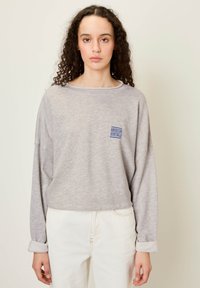 Siva majica z dolgimi rokavi in sproščenim krojem ter okroglih izrezom, z modrim logotipom "American Vintage" na prsih. Bele kavbojke s krajši ovratnikom.