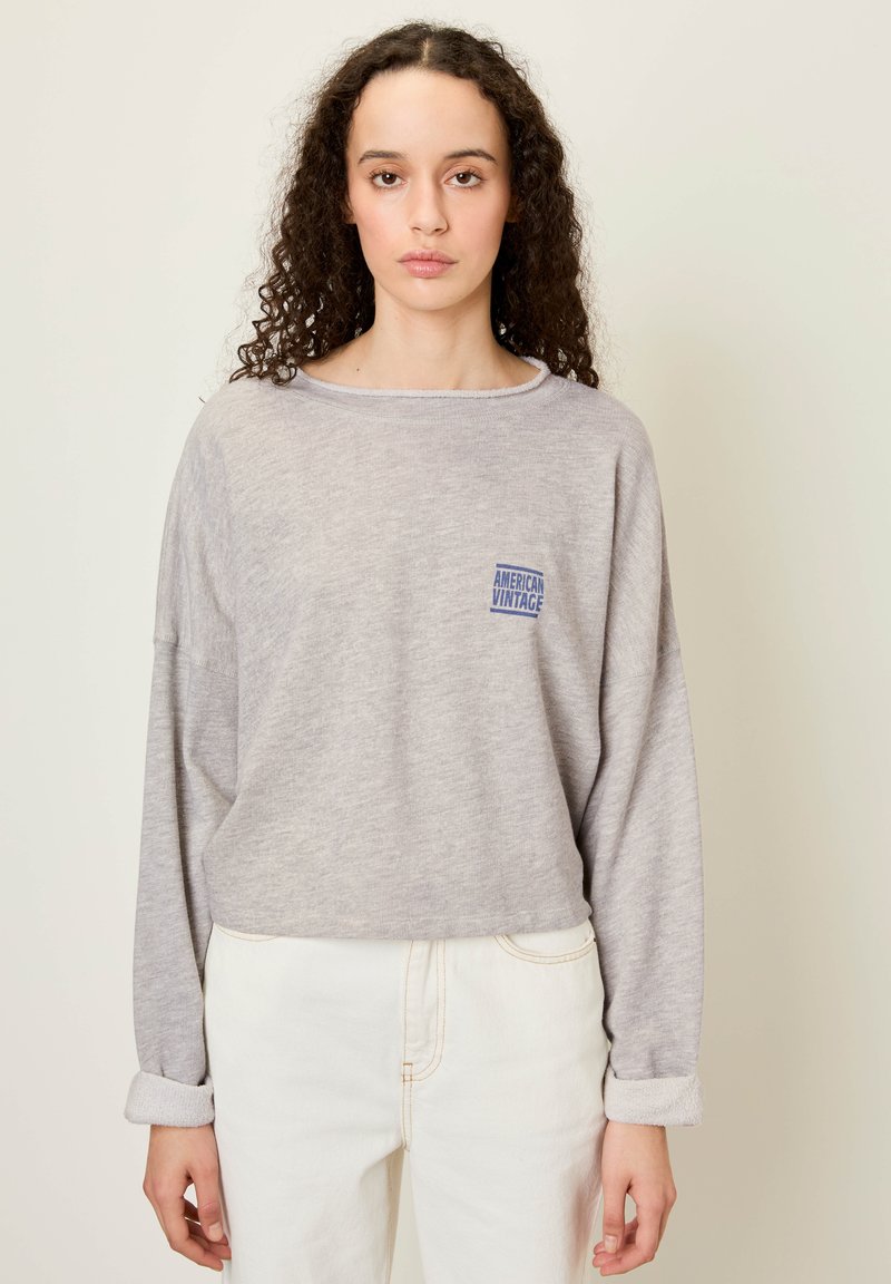 Siva majica z dolgimi rokavi in sproščenim krojem ter okroglih izrezom, z modrim logotipom "American Vintage" na prsih. Bele kavbojke s krajši ovratnikom.