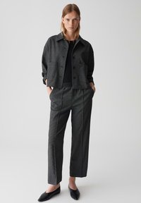 Person steht mit den Händen in den Taschen, trägt eine dunkelgraue karierte Jacke, passende Hose, ein schwarzes Oberteil und schwarze spitze Ballerinas vor einem weißen Hintergrund.
