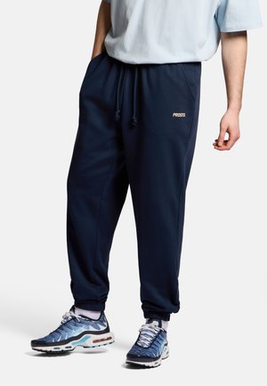 Mężczyzna noszący granatowe spodnie dresowe z beżowym logo „PROSTO”, białe skarpetki i niebieskie wzorzyste trampki, stojący na białym tle.