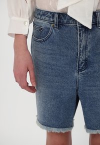 Bermuda in denim blu con orlo sfrangiato, dotata di un classico design a cinque tasche e accessori in ottone. Il tessuto presenta un aspetto scolorito e testurizzato.