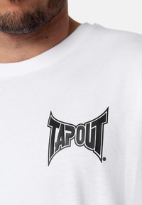 Bílý bavlněný tričko s výrazným černým logem "TAPOUT" na hrudi. Krátký rukáv, kulatý výstřih s hladkou texturou.