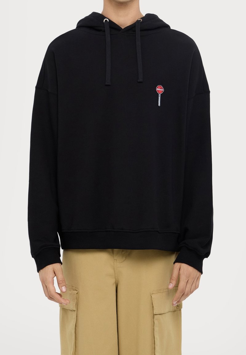 Personne portant un sweat à capuche noir avec cordons et un petit patch sucette rouge sur la poitrine, associé à un pantalon cargo beige.