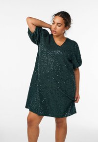 Abito verde con paillettes, scollatura a V e maniche a palloncino corte. Il tessuto presenta una superficie testurizzata decorata con paillettes luccicanti.