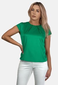 Zielona bluzka z krótkim rękawem o plisowanym dekolcie, wykonana z gładkiego materiału, w połączeniu z białymi spodniami, które mają luźny krój i prosty design.