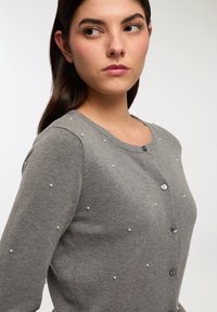 Cardigan grigio a maglia con scollo a giro, adornato con piccole perle e bottoni scuri lungo la parte anteriore per la chiusura.