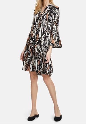 Frau trägt ein knielanges, langärmeliges Kleid mit braunen, schwarzen und weißen abstrakten Blattmustern und schwarzen, flachen Slip-On-Schuhen.