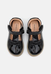 Zapatos de charol negro con forma redondeada, correa ajustable y acento de lazo negro texturizado en la parte delantera. Forro interior beige.