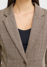 Blazer a righe marroni con colletto a rever, chiusura con un bottone e tessuto liscio, indossato sopra un top nero.