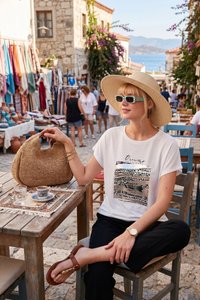 Frau mit Sonnenhut und Sonnenbrille sitzt an einem Tisch in einem Straßencafé mit einer geflochtenen Tasche, einer Kaffeetasse und einem steinernen Straßenmarkt mit Menschen im Hintergrund.