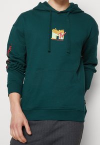 Sweat à capuche vert avec une poche avant, arborant un logo MTV jaune et rouge accompagné de frites. Poignets et ourlet côtelés, fabriqué dans un matériau doux.