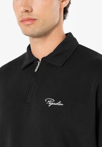 Sort polo skjorte med lynlås krave, lavet af blødt stof, har hvidt broderet "Pegador" logo på venstre bryst.