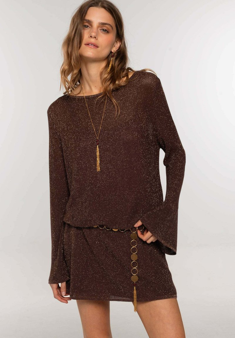 Top de punto marrón brillante con mangas anchas, superpuesto a una falda a juego con un cinturón de cadena con acentos circulares y un detalle de borlas.