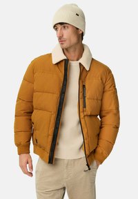 Bruine pufferjack met een crèmekleurige schapenvacht kraag, voorzien van zwarte ritsaccenten, gedragen over een beige gebreide trui. Een crèmekleurige beanie maakt de look af.