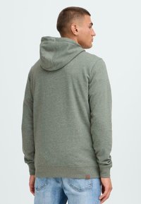 Hoodie u maslinastozelenoj tkanini, s opuštenim krojem, rebrastim rukavima i malom smešnom etiketom na rubu. Klasičan dizajn s mekanom teksturom.