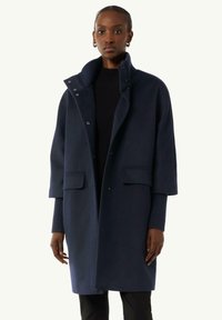 Marineblauer Wollmantel mit hohem Kragen, Druckknopfverschluss, zwei Fronttaschen und Ärmeln in Ellbogenlänge. Glatte Textur und taillierte Form.