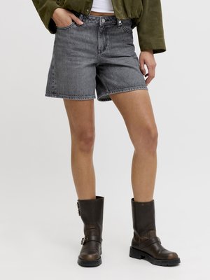 Osoba oblečená v sivých džínsových šortkách, olivovo zelenom kabáte, bielom krátkom tričku a tmavohnedých kožených topánkach po lýtka so sponami.