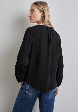 Schwarze, strukturierte Bluse mit langen, gerafften Ärmeln und rundem Halsausschnitt, mit einem dezenten Rückenschlitz, kombiniert mit blauen Jeans.