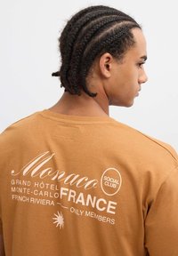 Camiseta de algodón marrón con texto blanco que incluye "Mónaco," "Grand Hôtel," y "Social Club." Mangas cortas y corte casual.