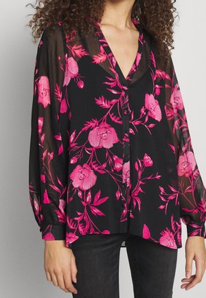 Läpinäkyvä musta blusa, jossa on kirkas vaaleanpunainen kukkakuosi, V-pääntie, pitkät hihat ja nappikiinnitys edessä, tyylitelty tummien farkkujen kanssa.