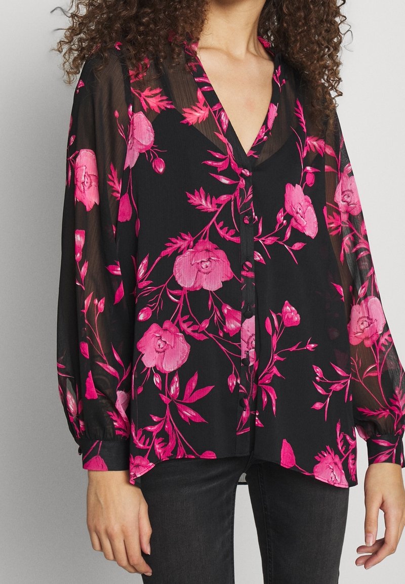 Blusa nera trasparente con vivace motivo floreale rosa, scollatura a V, maniche lunghe e chiusura con bottoni, abbinata a jeans scuri.
