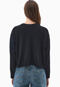 Schwarzer, kurz geschnittener Pullover mit geripptem Saum und Bündchen, mit rundem Halsausschnitt und Schulterakzenten. Getragen über hellblauen, abgenutzten Jeans.