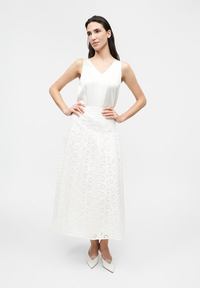 Γυναίκα ποζάρει με λευκό αμάνικο τοπ και μακριά λευκή φούστα με λουλουδάτο μοτίβο κοπής, φορώντας λευκές μυτερές γόβες.