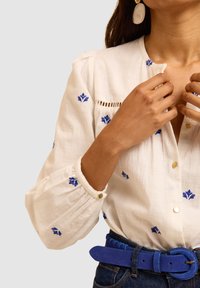Blusa di lino bianca con ricami floreali blu, scollatura tonda, chiusura con bottoni e maniche arricciate; abbinata a una cintura in camoscio blu.