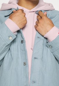 Chaqueta de mezclilla azul claro con botones a presión plateados, superpuesta a una sudadera rosa con cordón. Las manos están agarrando la chaqueta.