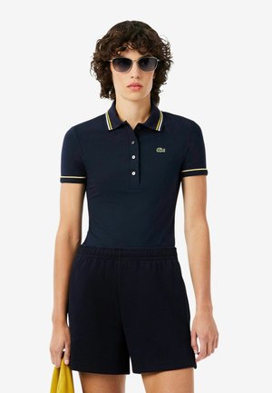 Mujer con polo negro con ribete amarillo, pantalones cortos negros, gafas de sol, sosteniendo un paño amarillo sobre fondo blanco.