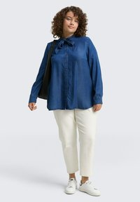 Denimowa koszula z guzikami w głębokim niebieskim kolorze z kokardą przy kołnierzy, zestawiona z kremowymi spodniami i białymi trampkami. Gładka faktura, luźny krój.