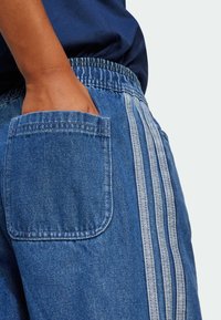 Τζιν σορτσάκι σε μπλε χρώμα με τσέπη ραμμένη, ελαστική μέση και τρεις κατακόρυφες γκρι ρίγες στο πλάι. Ορατή υφή υφάσματος.