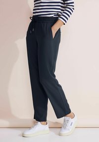 Donkergrijze joggerbroeken met een elastiche tailleband en een trekkoord. Voorzien van zijzakken en een taps toelopend ontwerp. Gecombineerd met witte sneakers.