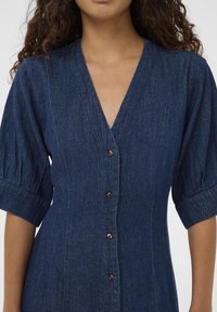Denim obleka z globokim V-izrezom in kratkimi naborki, z gumbi spredaj in prilegajočim se dizajnom. Temno modra barva in teksturirana podjet. 