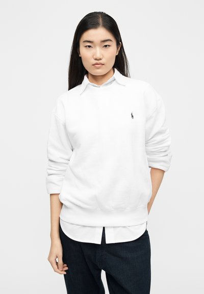 Γυναίκα με μακριά σκουρόχρωμα μαλλιά, φορώντας λευκό φούτερ πάνω από πουκάμισο με γιακά και σκούρο τζιν, όρθια με το ένα χέρι στην τσέπη.